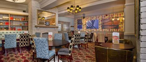 A hotel dining area with round tables, patterned chairs, a bookshelf, and a mirror.