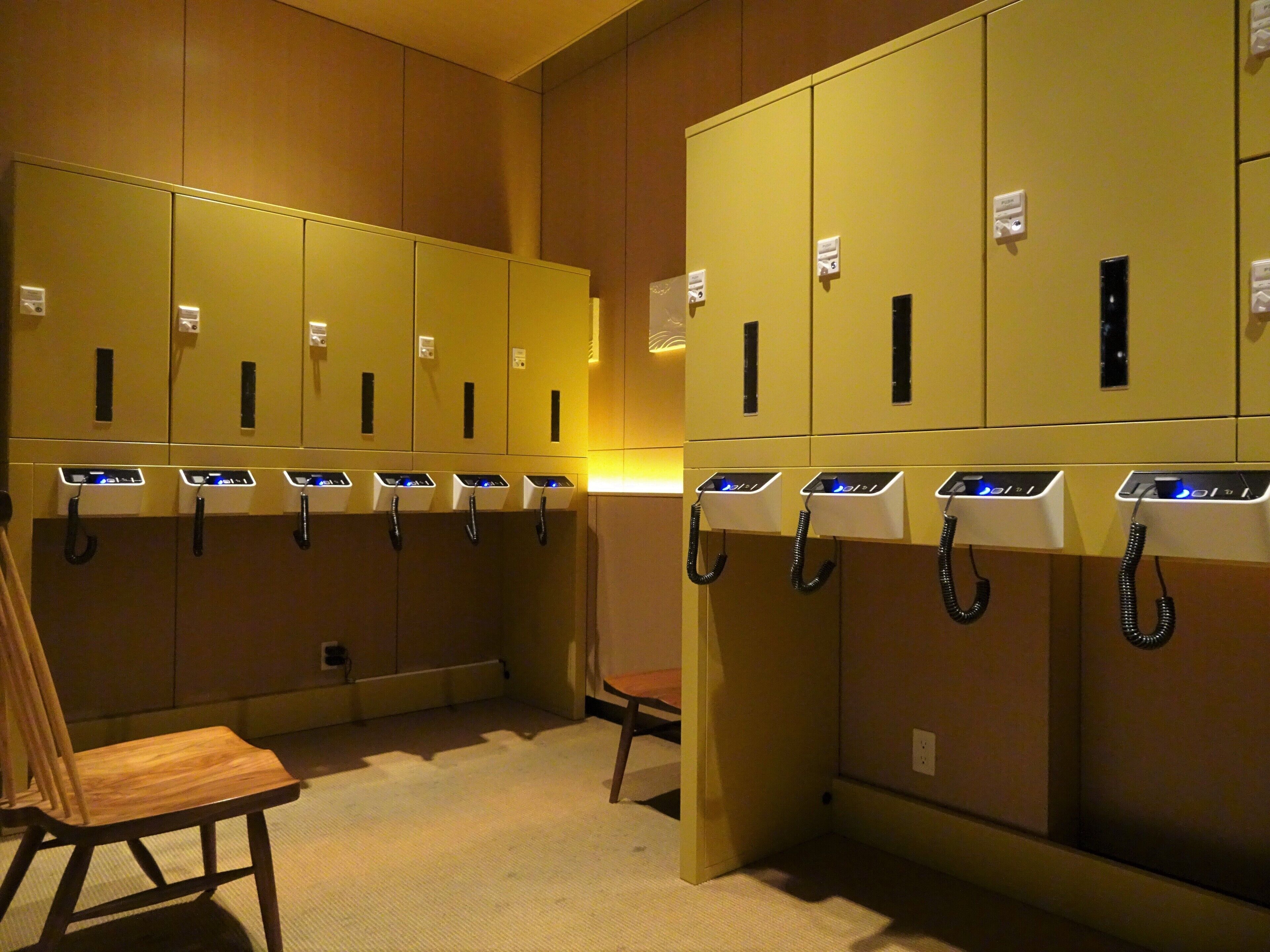 A row of lockers with charging stations, a wooden chair, and a small table in a well-lit room.