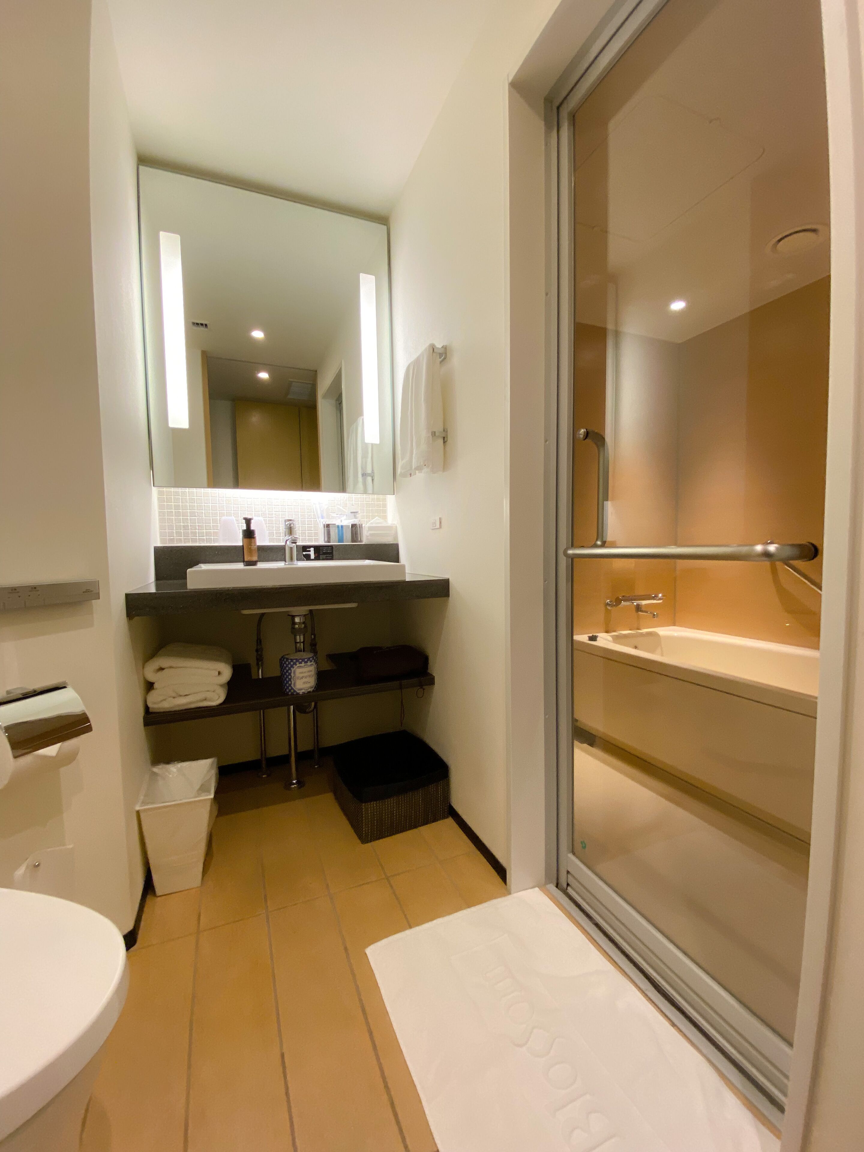 Un baño moderno con un espejo grande, una bañera y un inodoro. Hay un perchero y un estante con artículos de aseo.