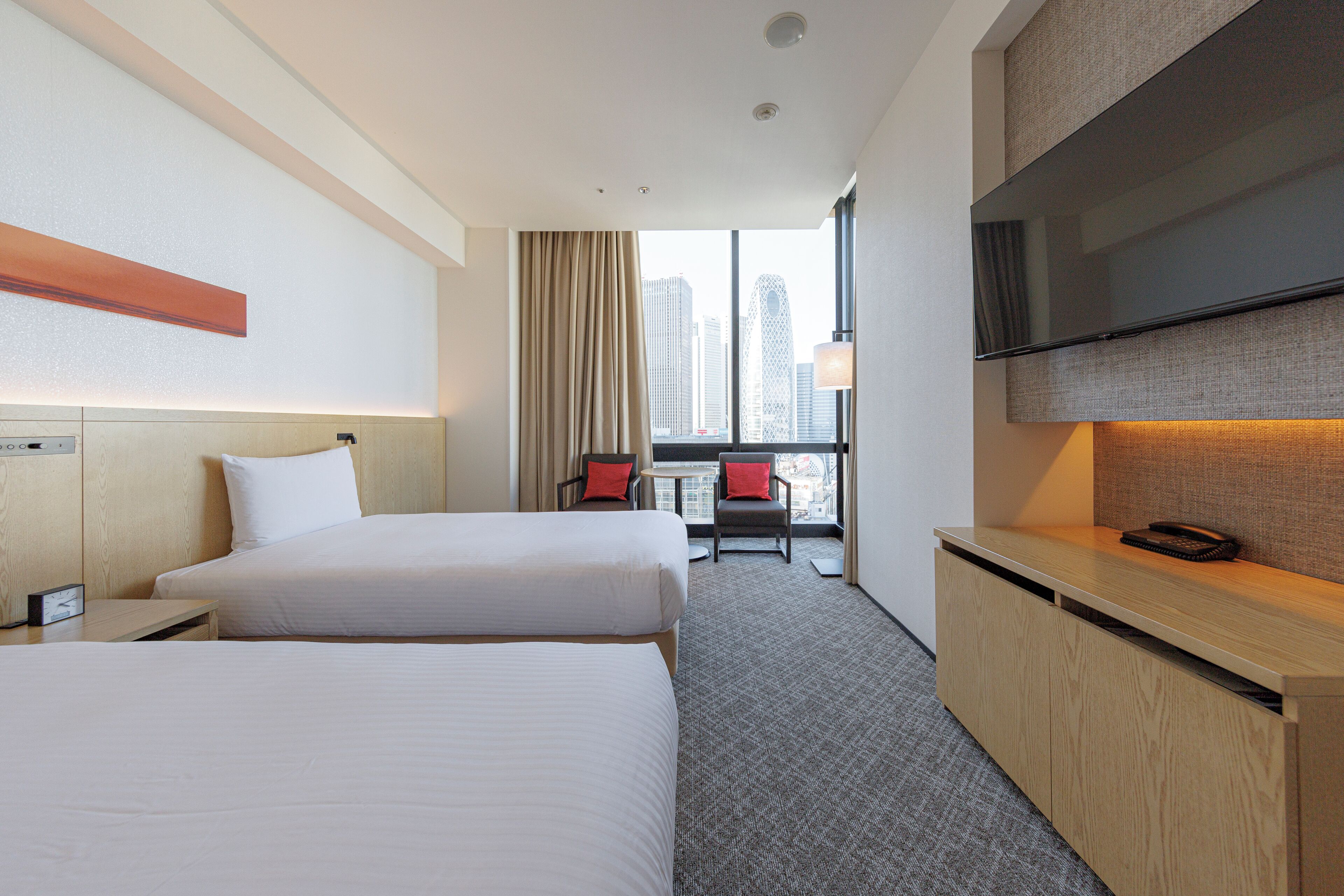 A hotel room with two beds, a desk, a TV, and a window with a city view.