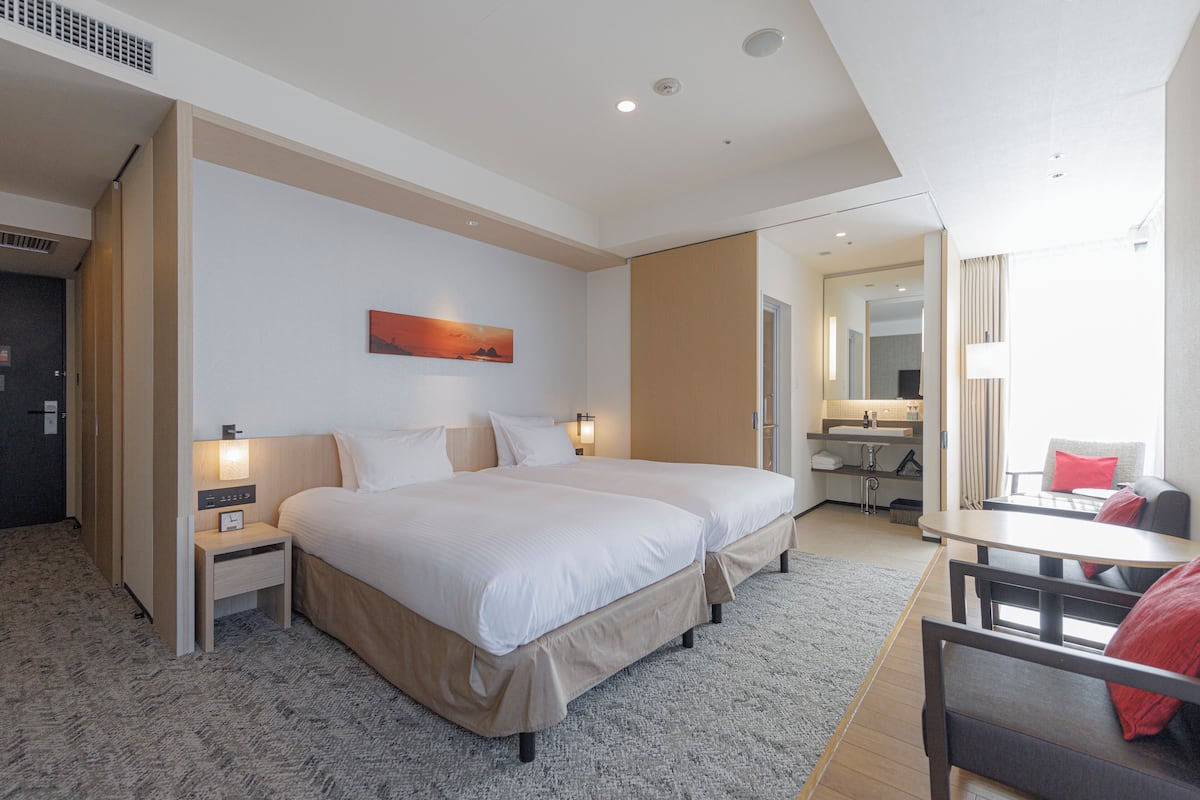 A hotel room with two beds, a desk, a chair, a nightstand, a lamp, a painting, and a bathroom visible through an open door.