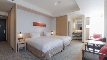 A hotel room with two beds, a desk, a chair, a nightstand, a lamp, a painting, and a bathroom visible through an open door.