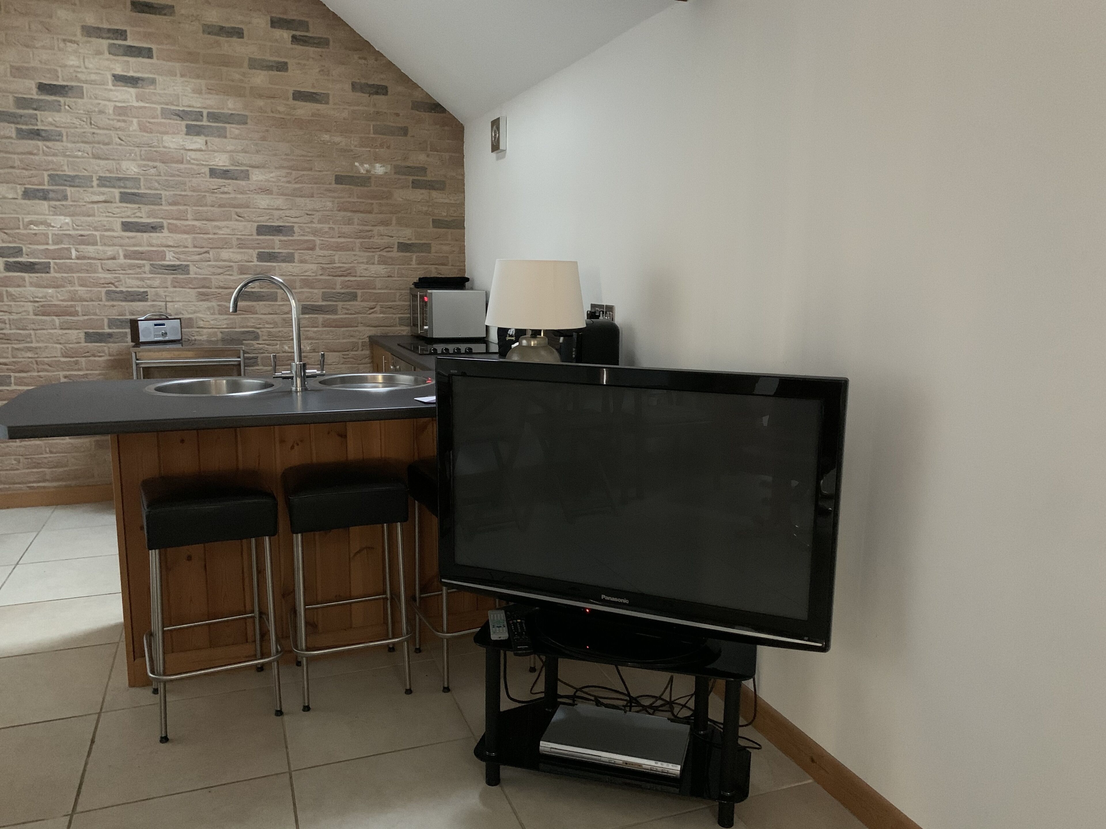 A compact kitchen with a built-in sink, a toaster, and a microwave. There are two bar stools and a flat-screen TV on a stand.