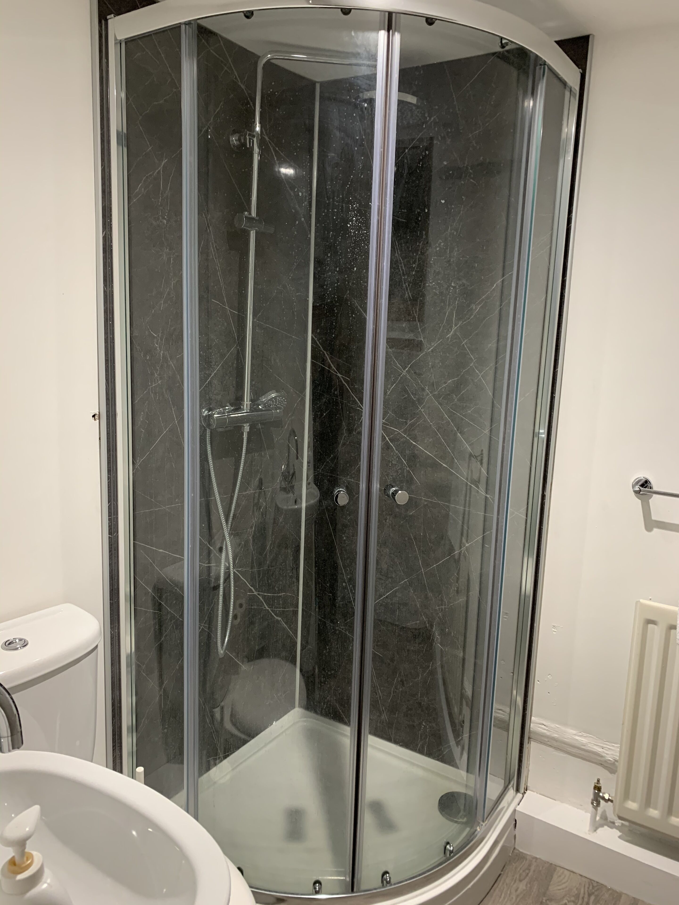A corner shower with a glass door, a white sink, and a white radiator.