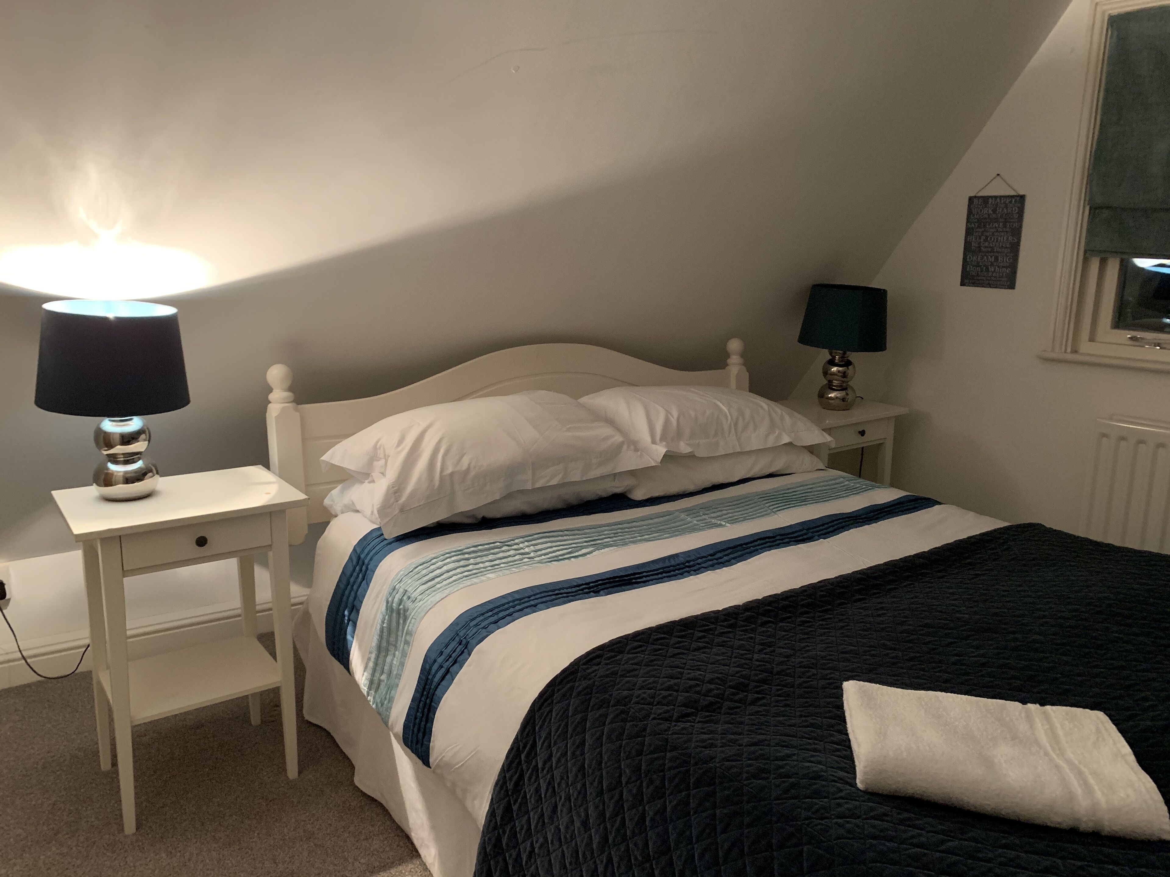 A bedroom with a bed, two bedside tables, a lamp on each table, a quilted bedspread, and a wall with a framed quote.