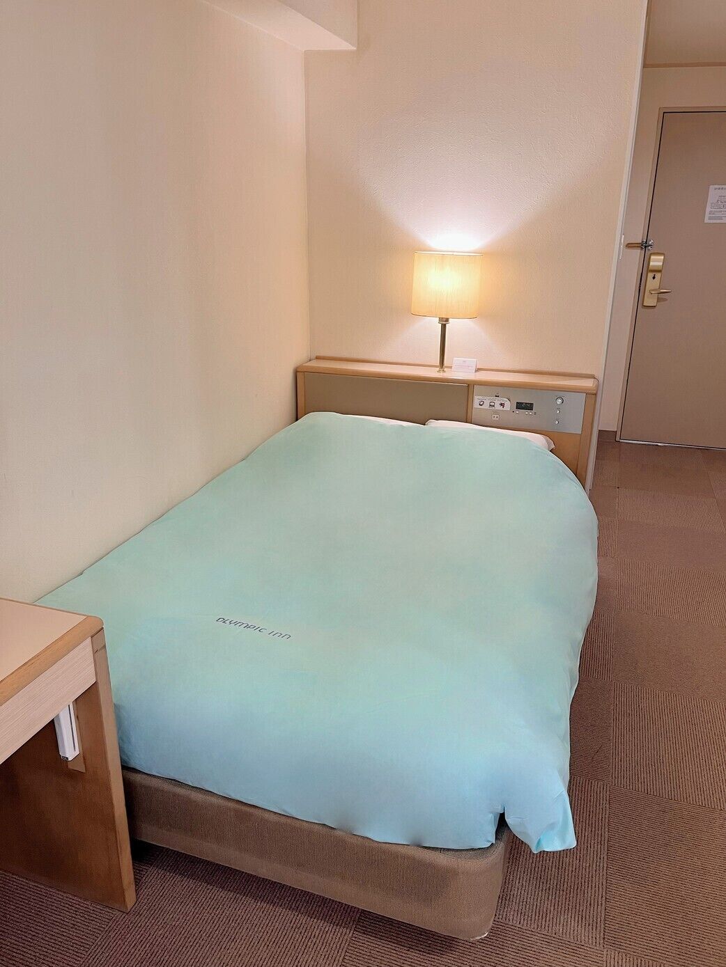 A hotel room with a bed, a bedside table, a lamp, and a door.