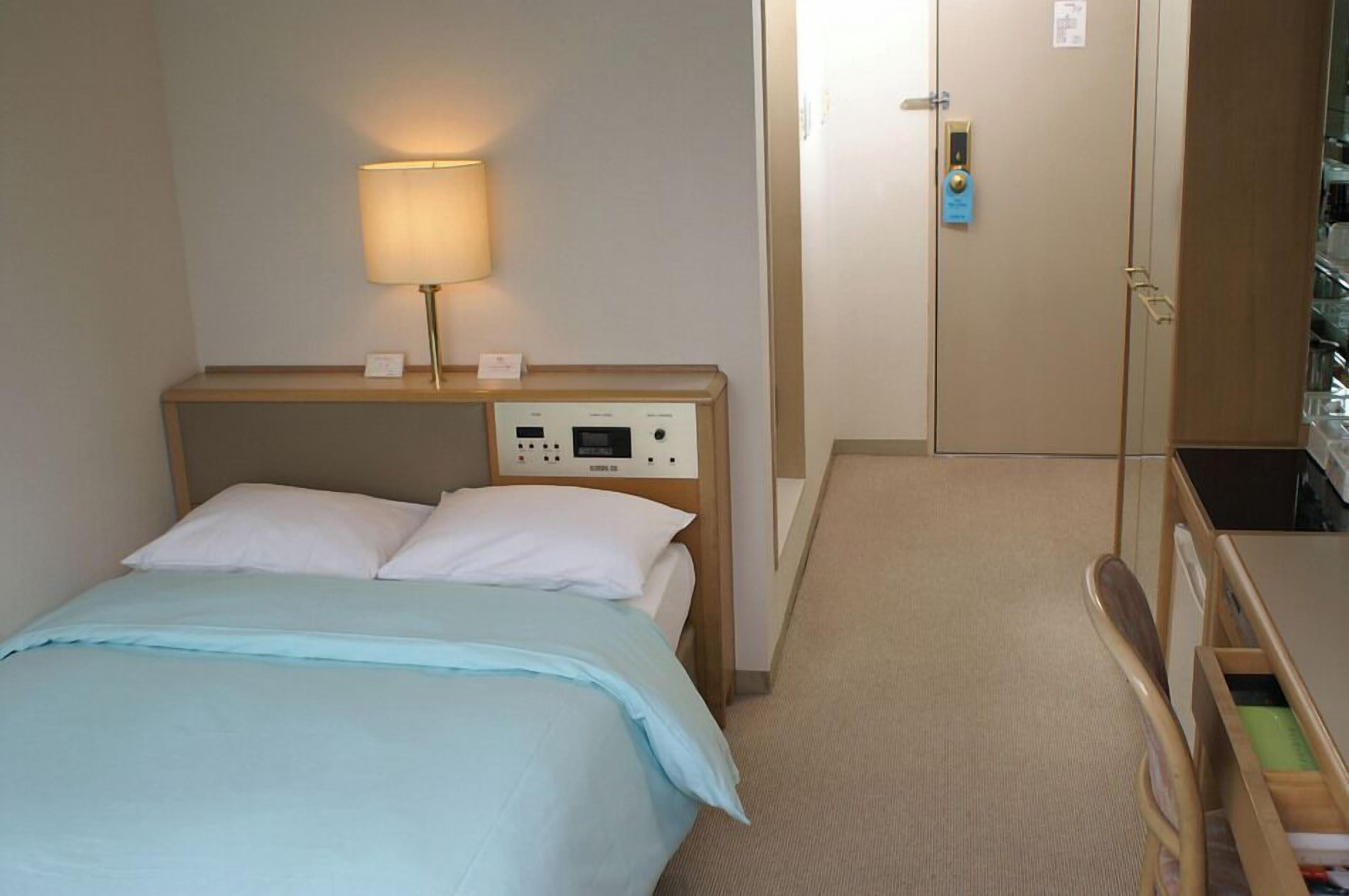 A hotel room with a bed, bedside lamp, control panel, wardrobe, and a small desk with drawers.