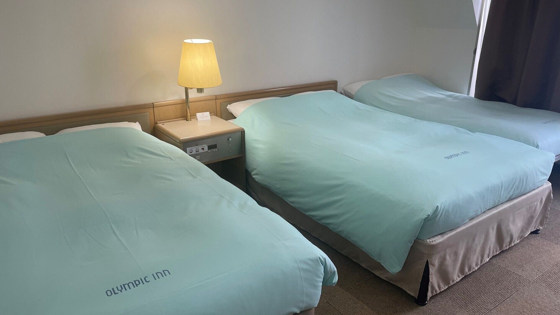 A hotel room with two beds, a bedside table with a lamp, and a control panel.
