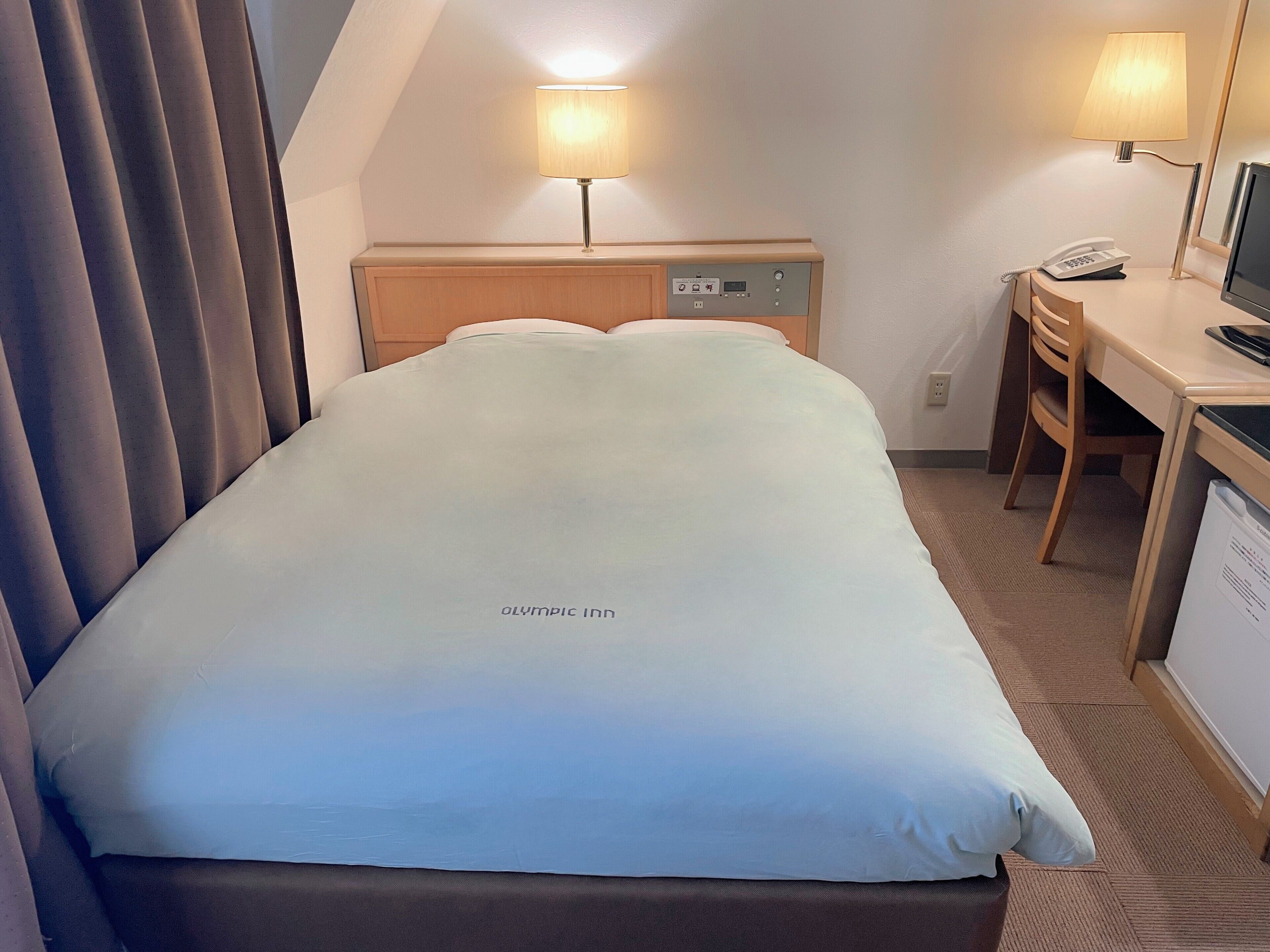 A hotel room with a bed, a desk, a chair, a lamp, a phone, and a television.