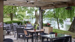 An outdoor patio with tables and chairs, overlooking a body of water with trees and boats. - The Rose Revived By Chef and Brewer Collection (Witney)