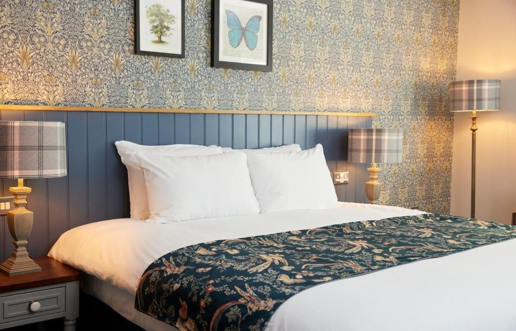 A neatly made bed with white linens, a dark patterned throw, and a blue headboard. There are two bedside lamps with plaid shades and a framed picture on the wall.