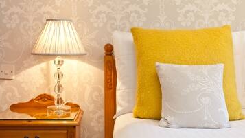 A neatly made bed with a white and yellow pillow set, a wooden nightstand with a lamp, and a patterned wallpaper background.