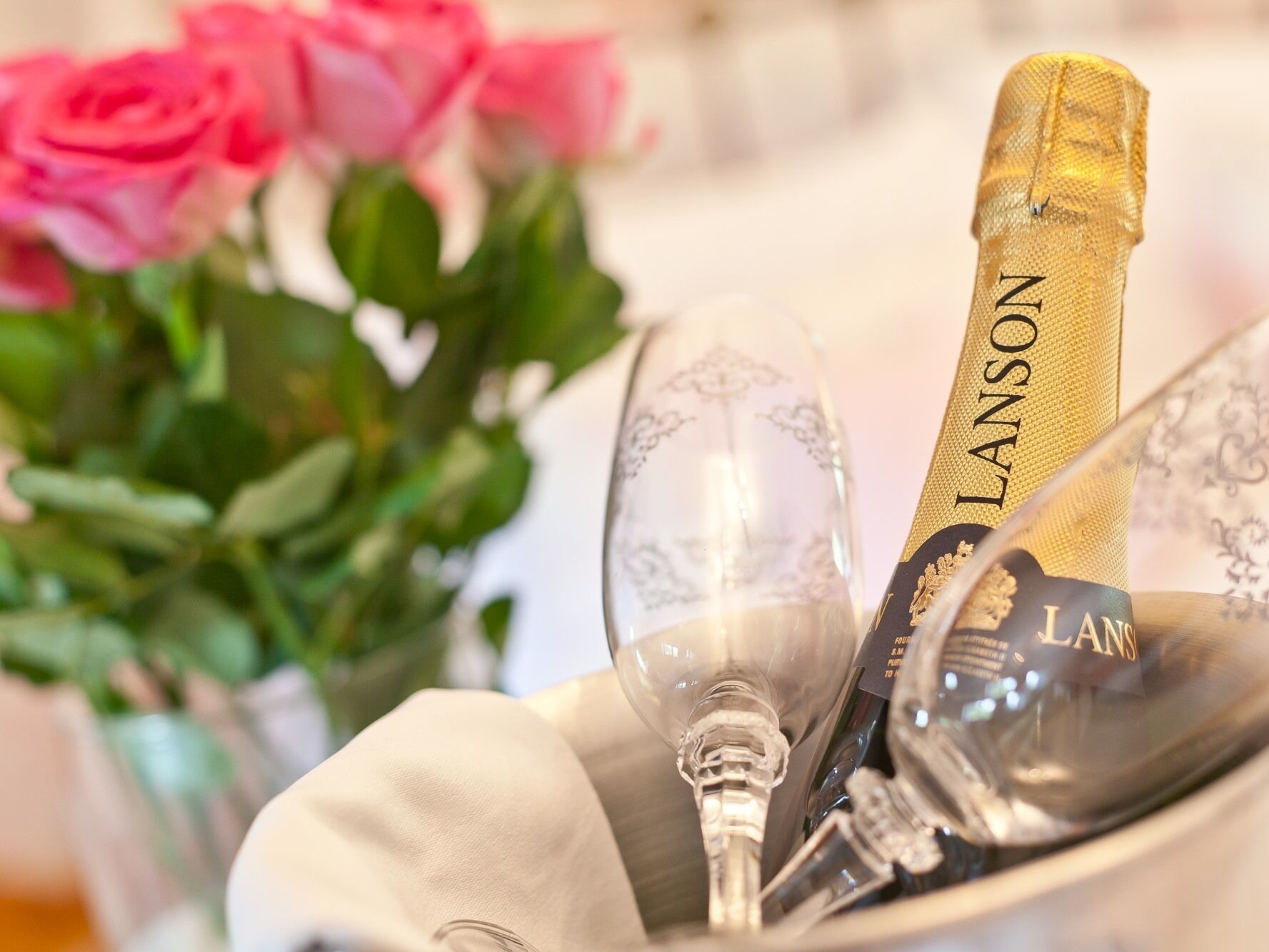 A champagne bottle labeled 'Lanson' and two clear wine glasses in a decorative setting with pink roses.