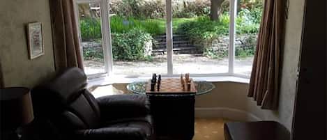 A room with a black leather sofa, a small round table with a chess set, and a large window with curtains.
