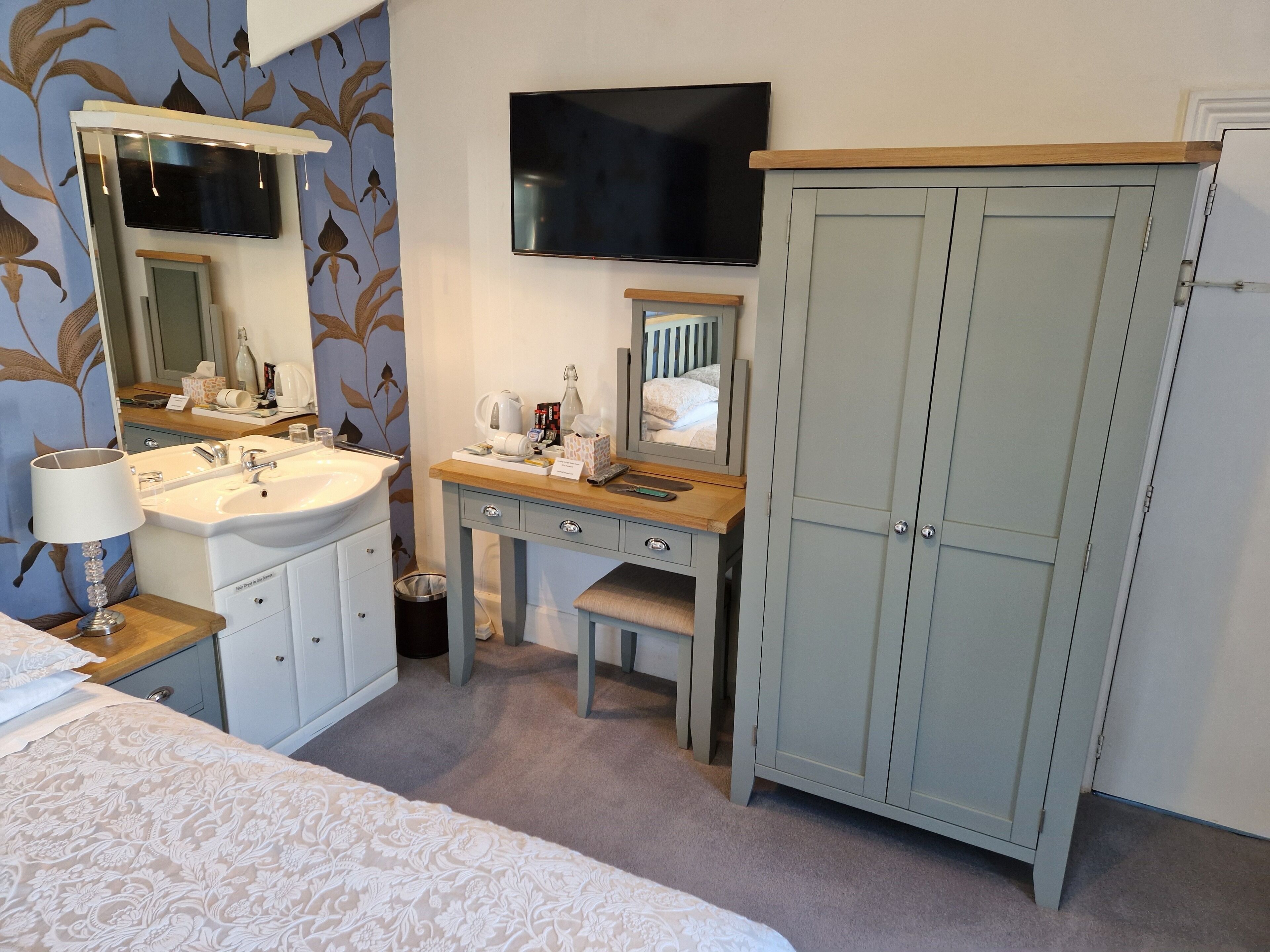 A hotel room with a bed, a bathroom with a sink, mirror, and toiletries, a TV, and a wardrobe.