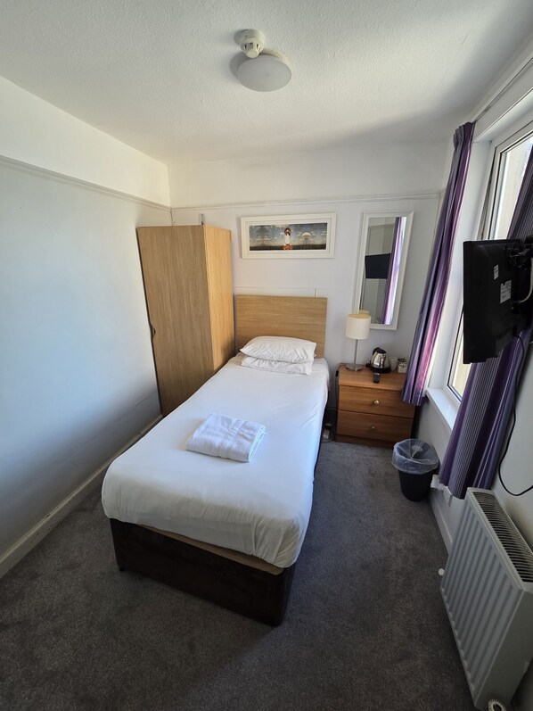 Hotelkamer met een bed, een houten garderobe, een nachtkastje, een prullenbak, een verwarming, een televisie en een raam met gordijnen.