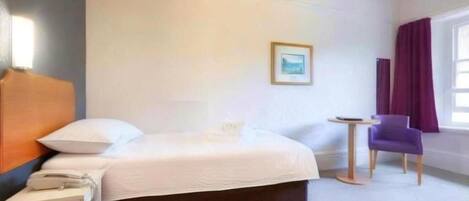 A hotel room with a bed, a bedside table with a phone, a small round table, a purple chair, and a window with curtains.