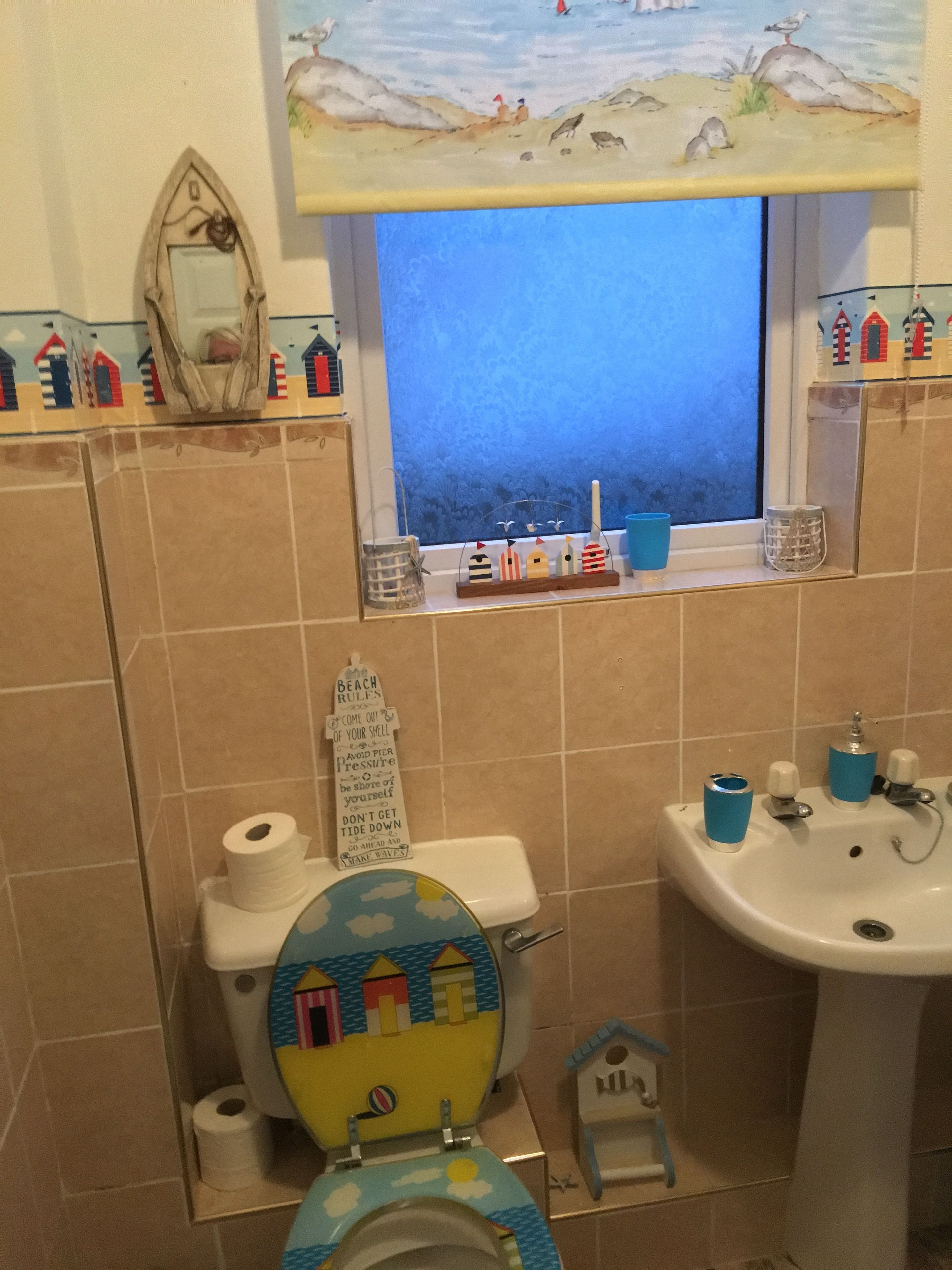 A bathroom with a toilet decorated with a beach scene, a window with a view of the sea, and a sink with blue cups.