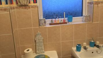 A bathroom with a toilet decorated with a beach scene, a window with a view of the sea, and a sink with blue cups.