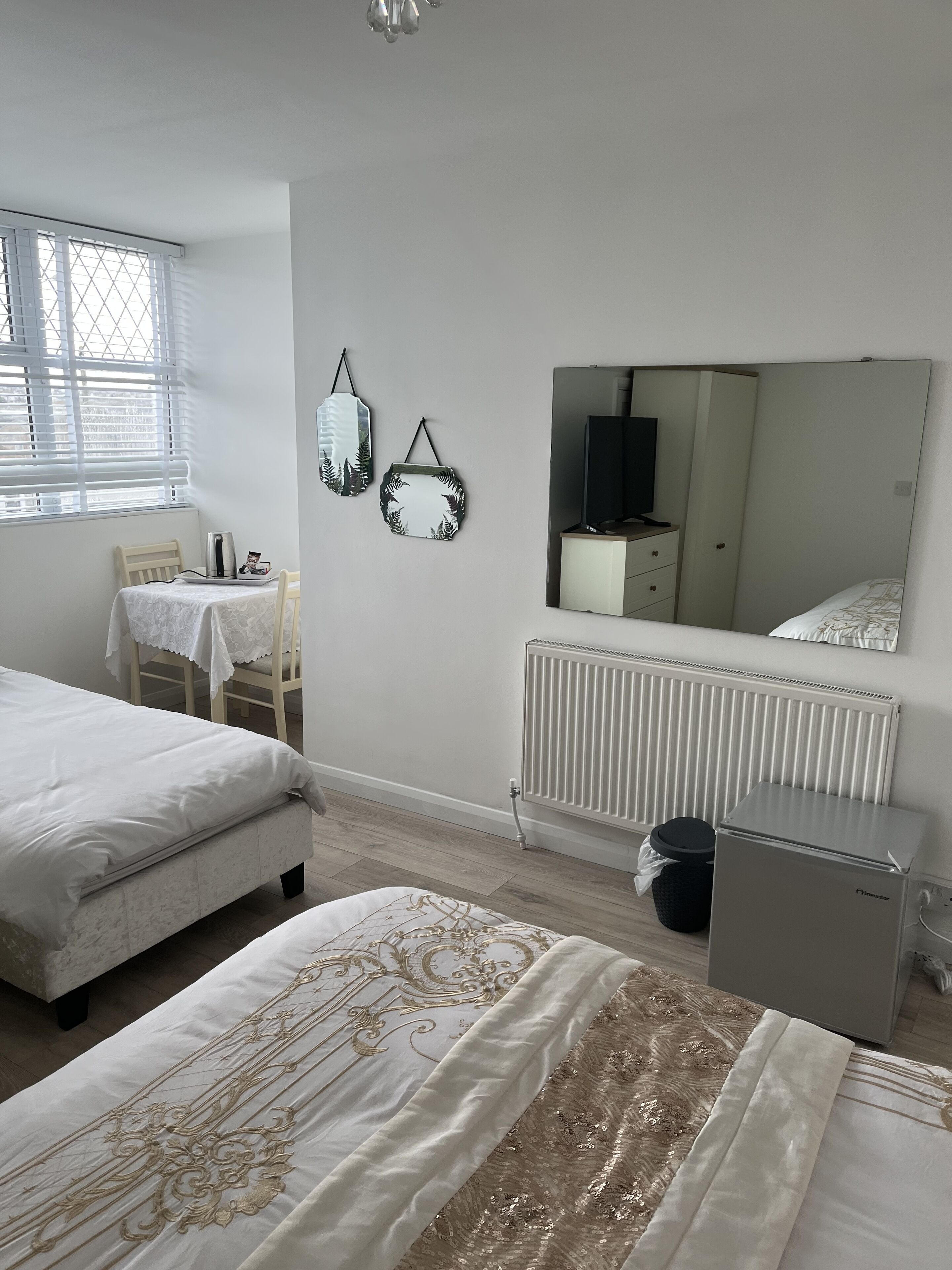 A bedroom with two beds, a radiator, a small refrigerator, a table with a chair, and a window with blinds.