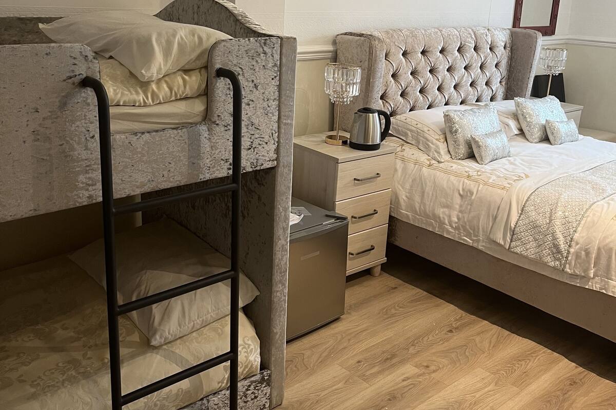 A bedroom with a bunk bed, a separate bed with a tufted headboard, a bedside table with a lamp, and a small cabinet.