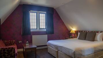 A bedroom with a sloped ceiling, a bed with white linens, a small table, a chair, and a window with curtains.