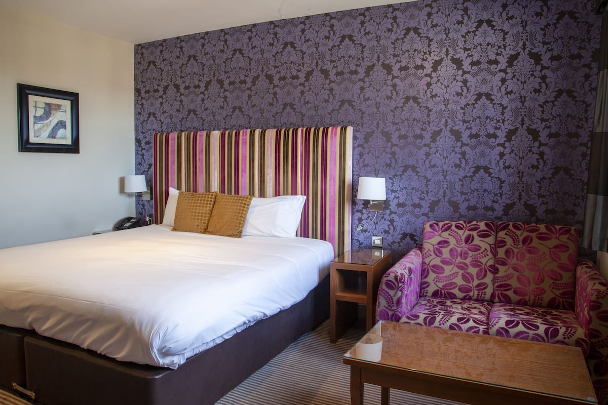 Une chambre d’hôtel avec un grand lit, un canapé, une table de chevet et un mur recouvert d’un papier peint à motifs.