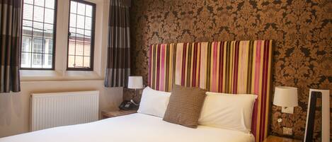 A hotel room with a large bed, a window with curtains, a radiator, a bedside table with a lamp, and a mirror.
