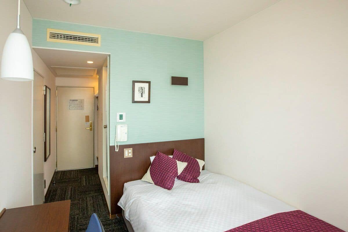 Een hotelkamer met een bed, een bureau, een stoel, een deur en een aan de muur bevestigd telefoon.