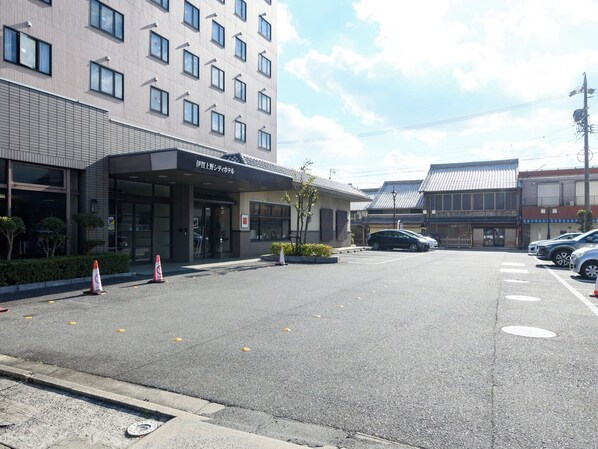 Entrada de un hotel con fachada moderna, un estacionamiento y edificios tradicionales al fondo.