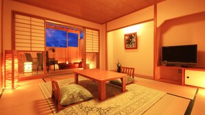 Une pièce de style japonais traditionnel, avec un sol recouvert de tatamis, une basse table en bois, un téléviseur et des portes coulissantes donnant sur une autre pièce.