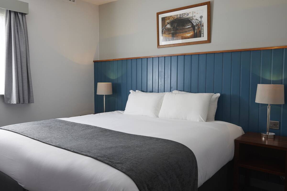 Une chambre d’hôtel avec un grand lit, deux tables de chevet avec des lampes, un tableau encadré au mur et une fenêtre avec des rideaux.