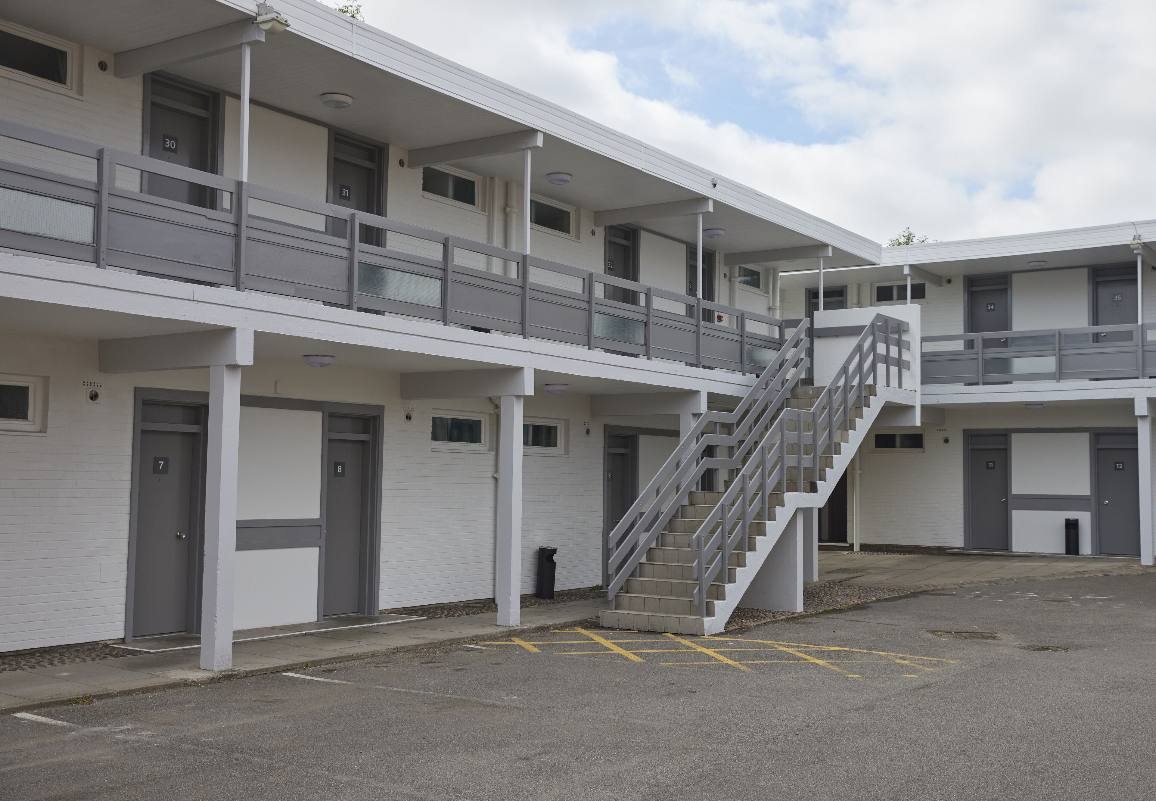 Un motel de varios pisos con una escalera central, habitaciones individuales numeradas del 7 al 12, y un área de estacionamiento con espacios delimitados.