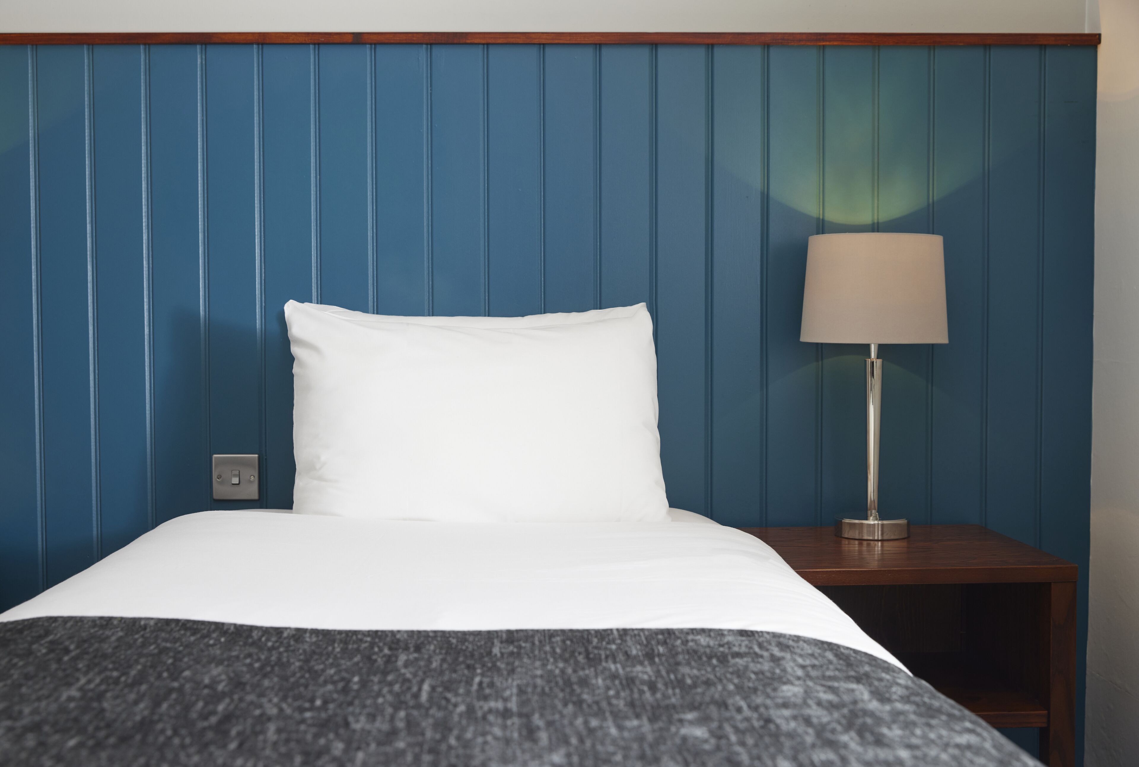 Una cama bien hecha con sábanas blancas, un plaid gris oscuro y una mesita de noche de madera con una lámpara.