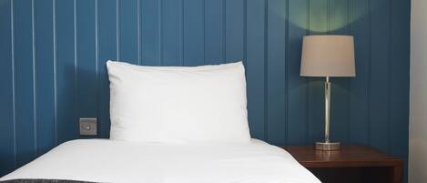 Una cama bien hecha con sábanas blancas, un plaid gris oscuro y una mesita de noche de madera con una lámpara.
