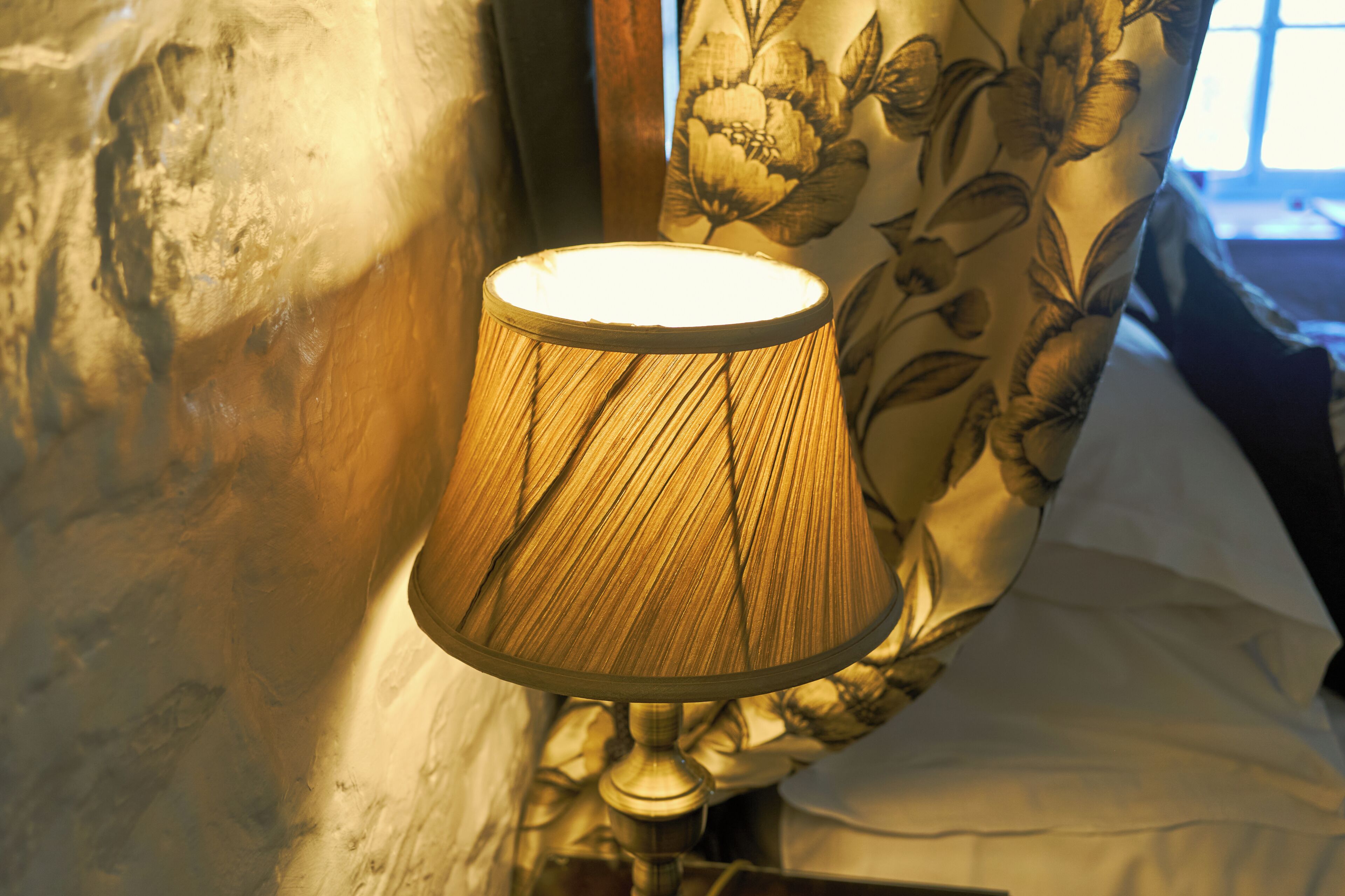 A lamp with a patterned shade, a floral curtain, and a bed with white sheets.