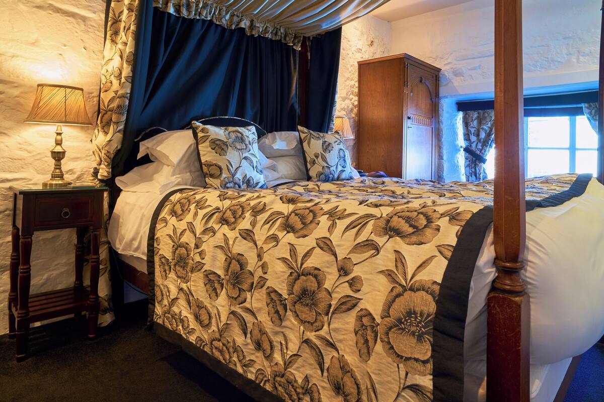 A four-poster bed with floral bedding, a nightstand with a lamp, and a wooden wardrobe in a room with stone walls.
