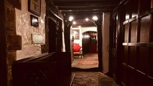 Un estrecho pasillo con paredes de piedra y puertas de madera, que conduce a una habitación con una silla roja y un cuadro enmarcado en la pared.