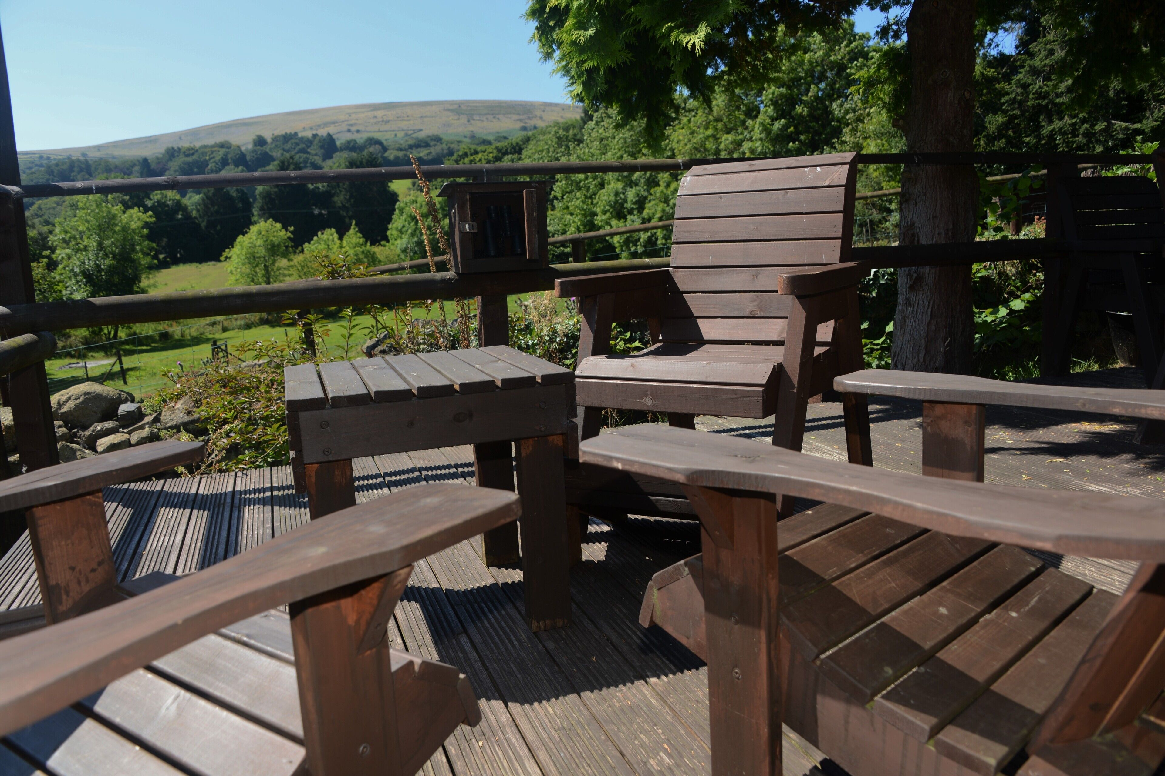 A wooden deck with benches and a table, overlooking a green landscape with trees and hills.