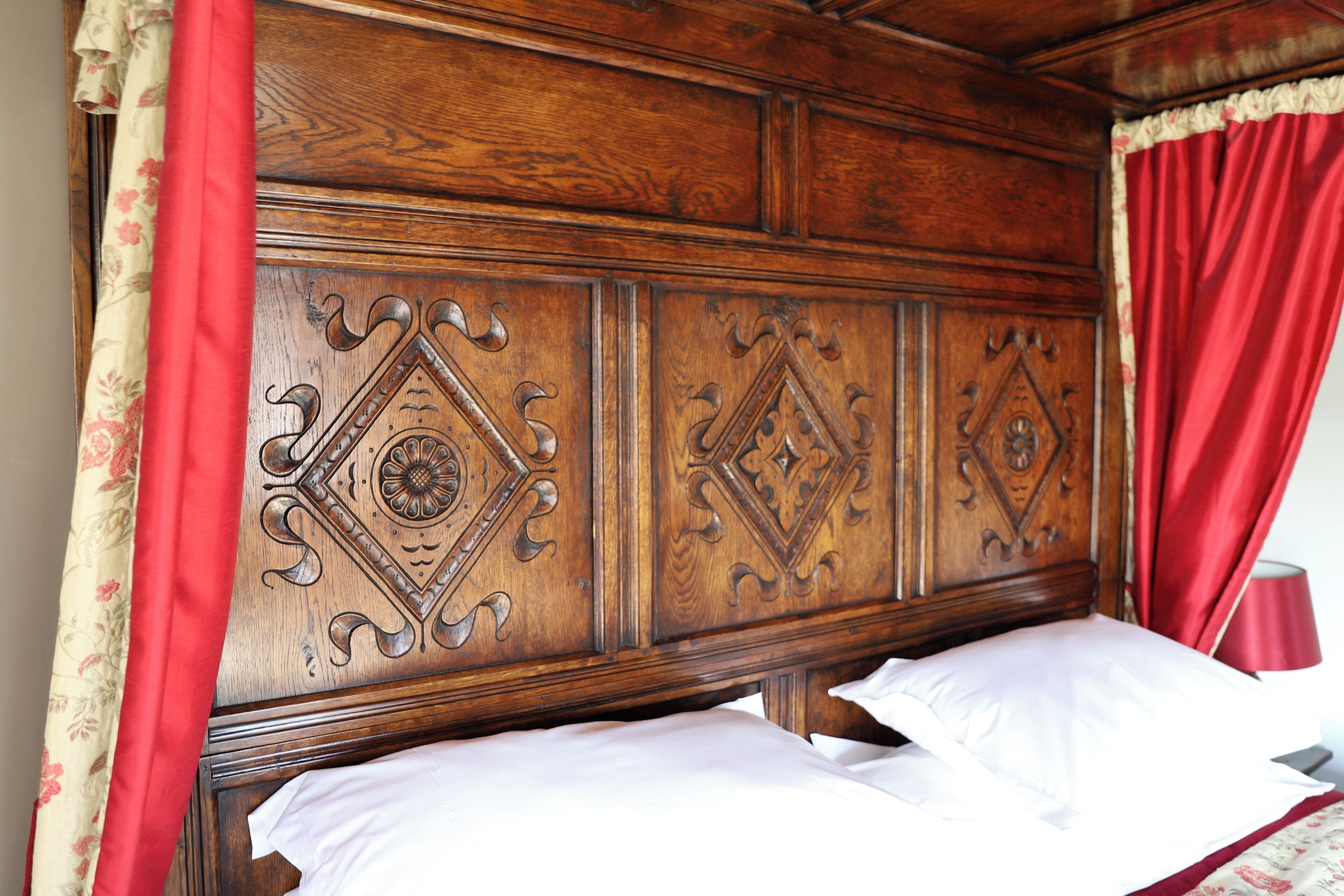 Cabecera de madera con intrincados grabados, cama con sábanas blancas y cortinas rojas.