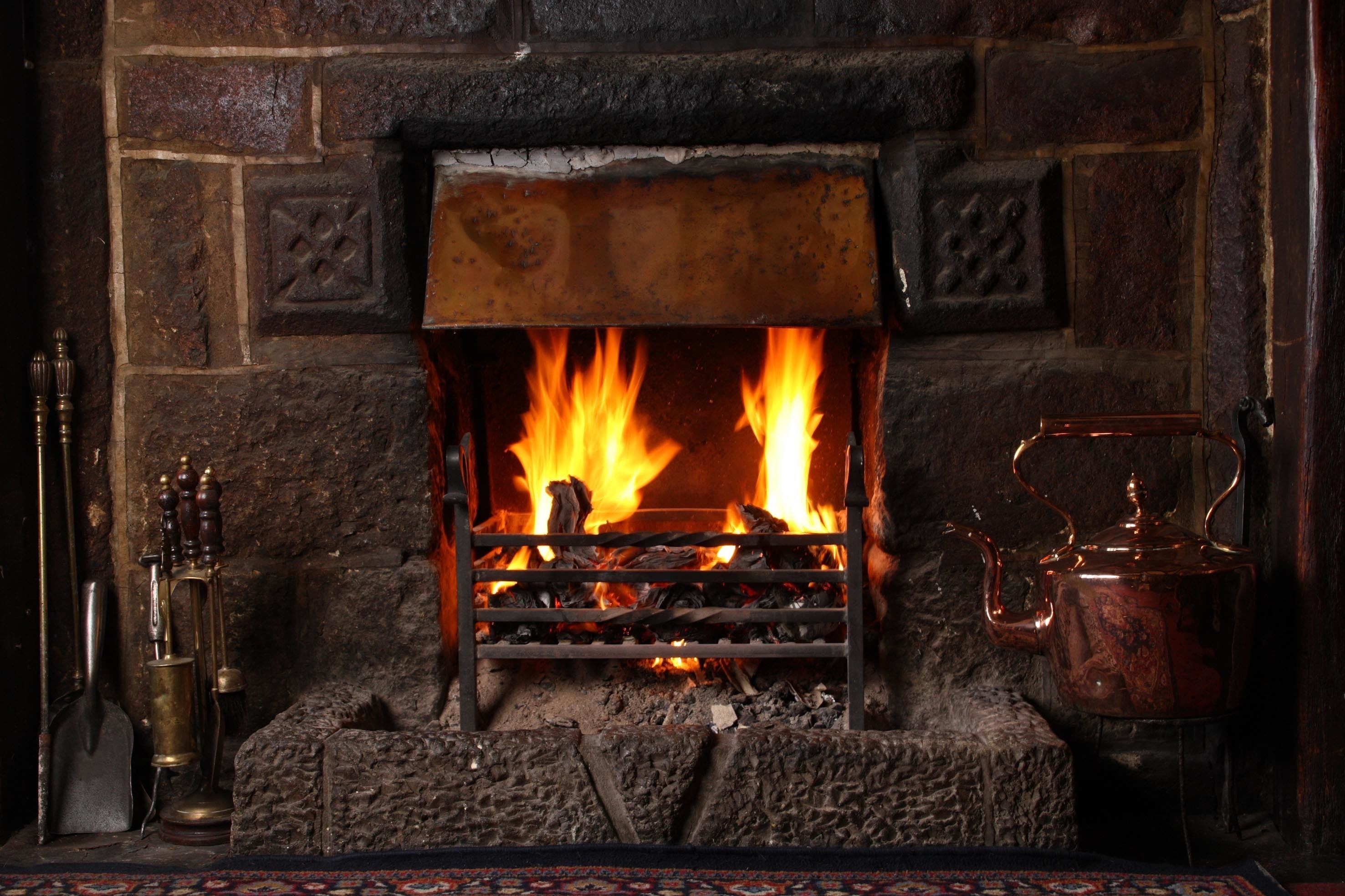 A fireplace with a metal grate, burning logs, and a copper kettle on a mantel.