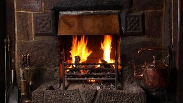 A fireplace with a metal grate, burning logs, and a copper kettle on a mantel.