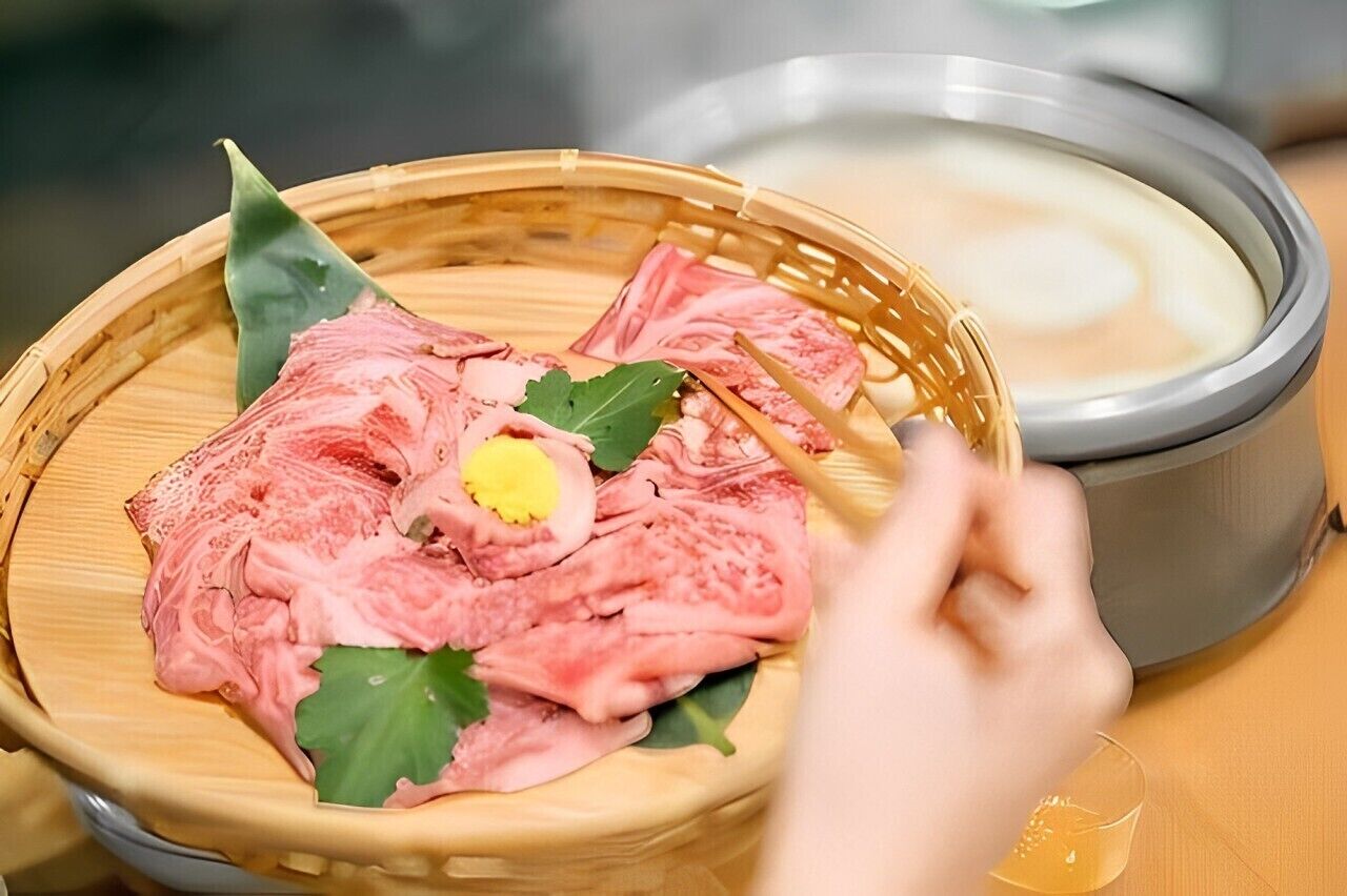 A hand holding chopsticks over a bamboo steamer containing sliced meat and a boiled egg, with a bowl of soup in the background.