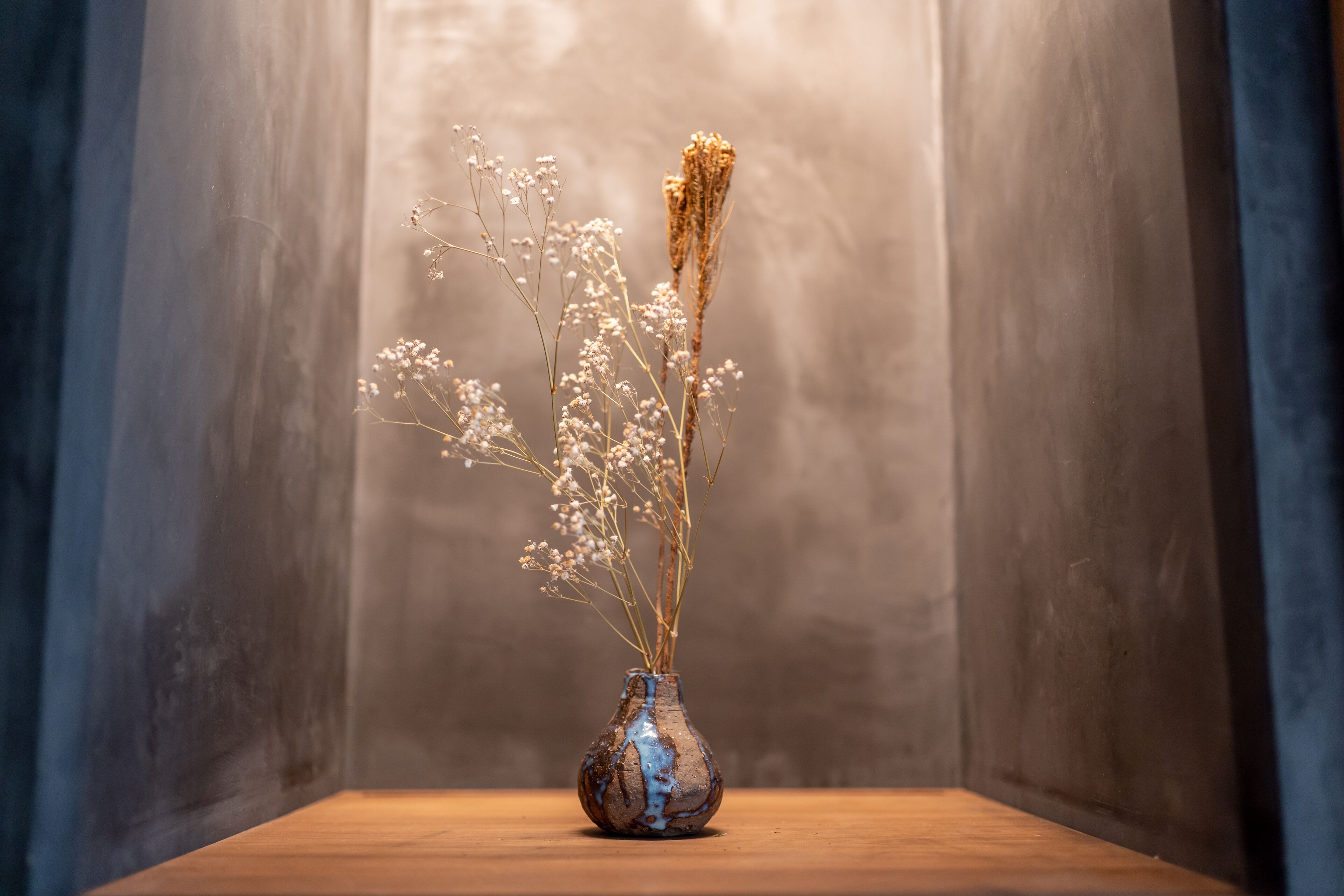Een vaas met gedroogde bloemen op een houten oppervlak, voor een structuurmuur.