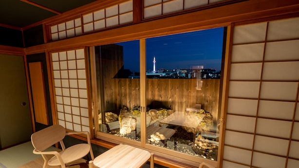 Ein traditioneller japanischer Raum mit Tatami-Bodenbelag, einem niedrigen Tisch und zwei Stühlen. Ein großes Fenster bietet Blick auf eine nächtliche Stadtlandschaft und einen Turm.