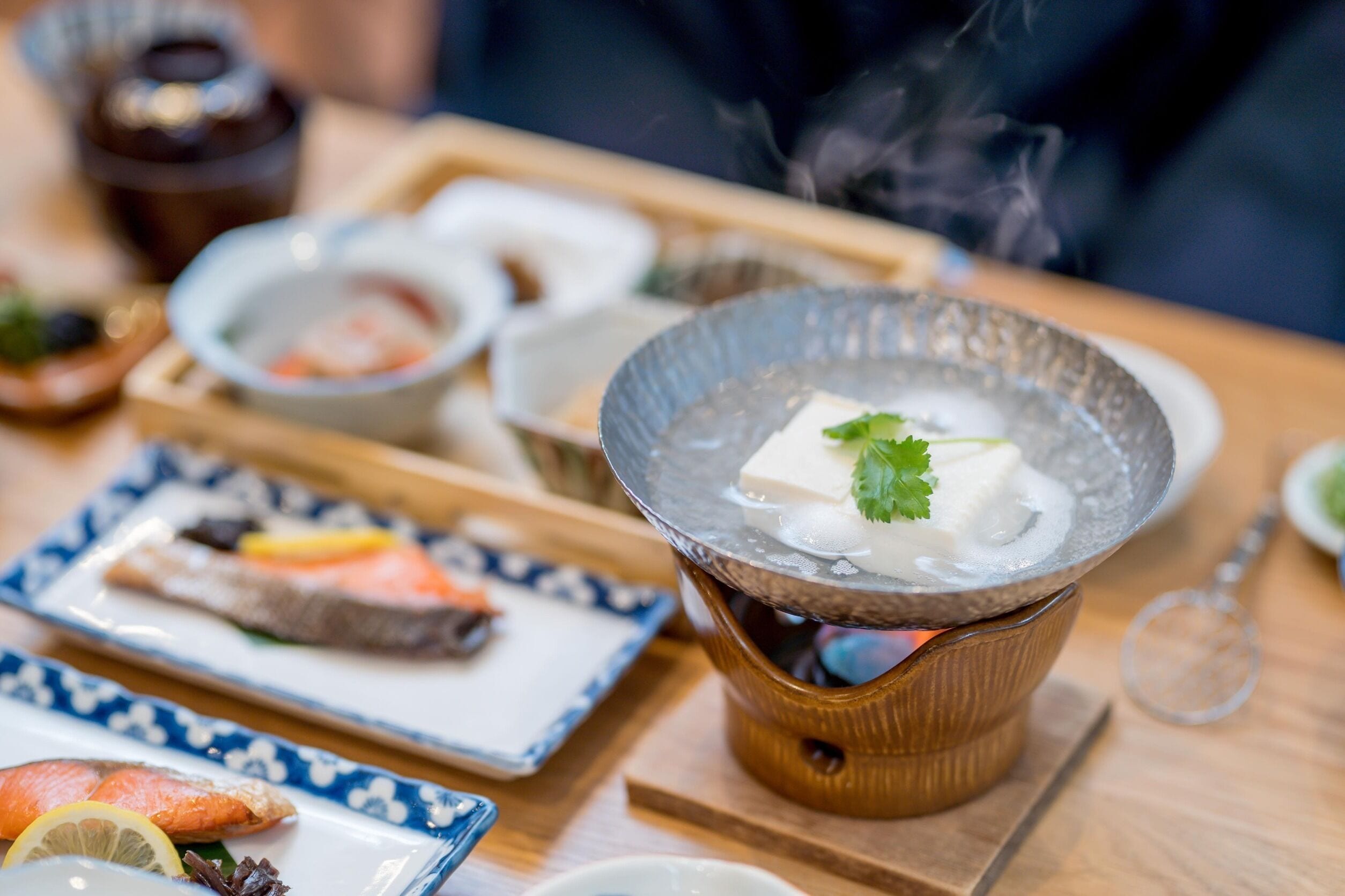 A hot pot setup with tofu and vegetables, surrounded by various dishes on a wooden table.