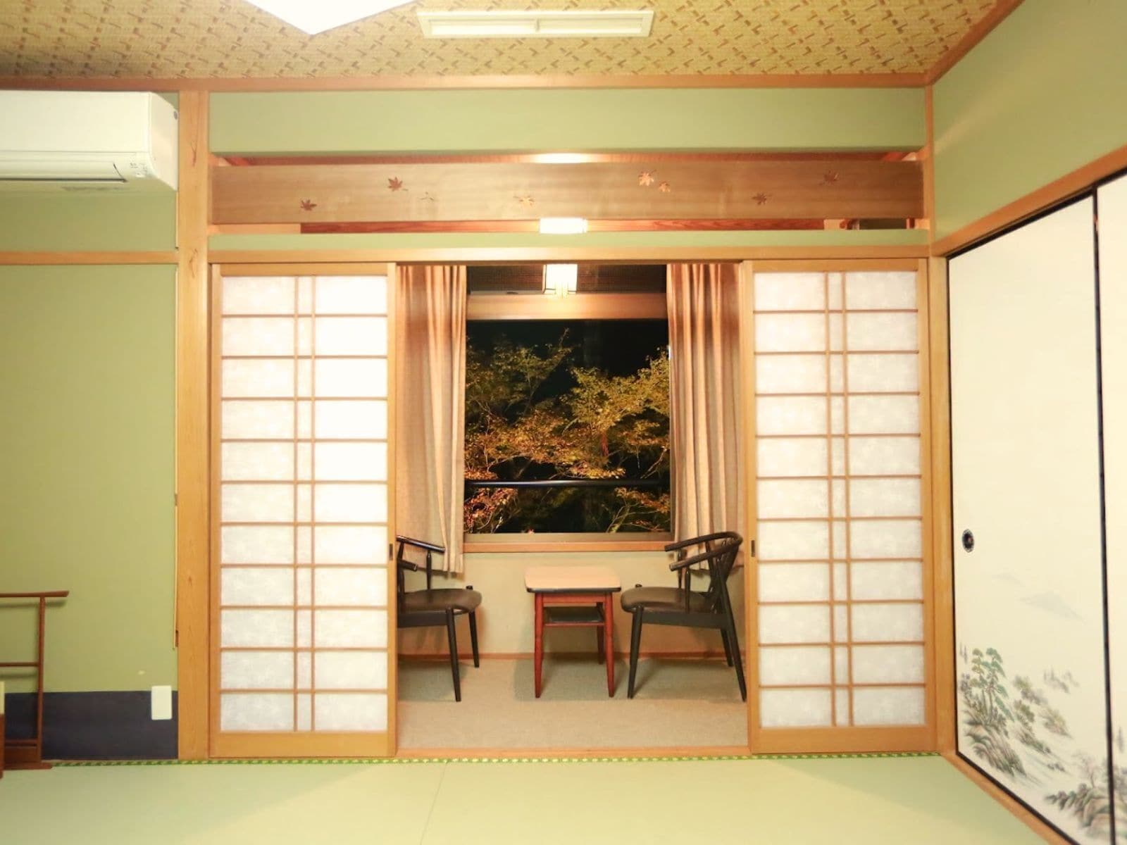 A traditional Japanese room with tatami flooring, a low table, two chairs, and a view of trees through shoji screens.