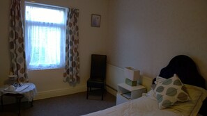 Un dormitorio con cama, una silla, una mesita con bandeja, un radiador y una ventana con cortinas.