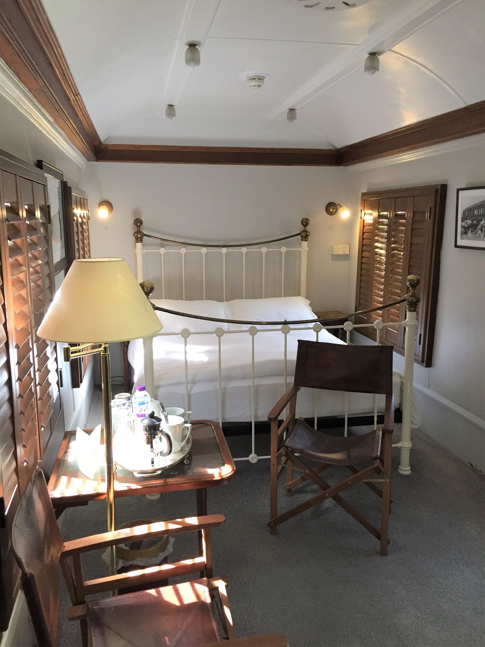 A hotel room with a single bed, a wooden chair, a table with a lamp, and a tray with a cup and saucer.