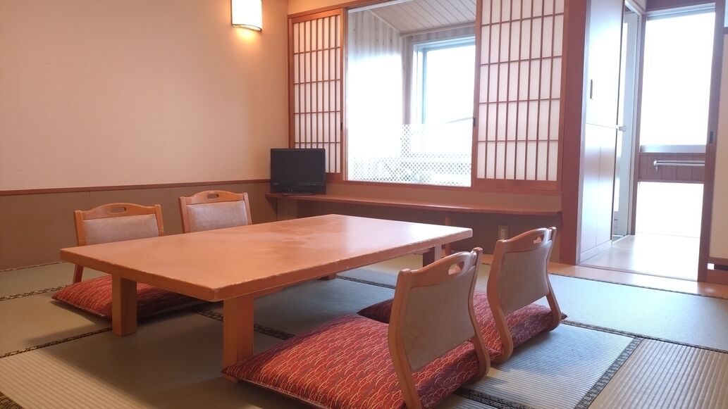 A traditional Japanese room with tatami flooring, a low table, four chairs, and a television.